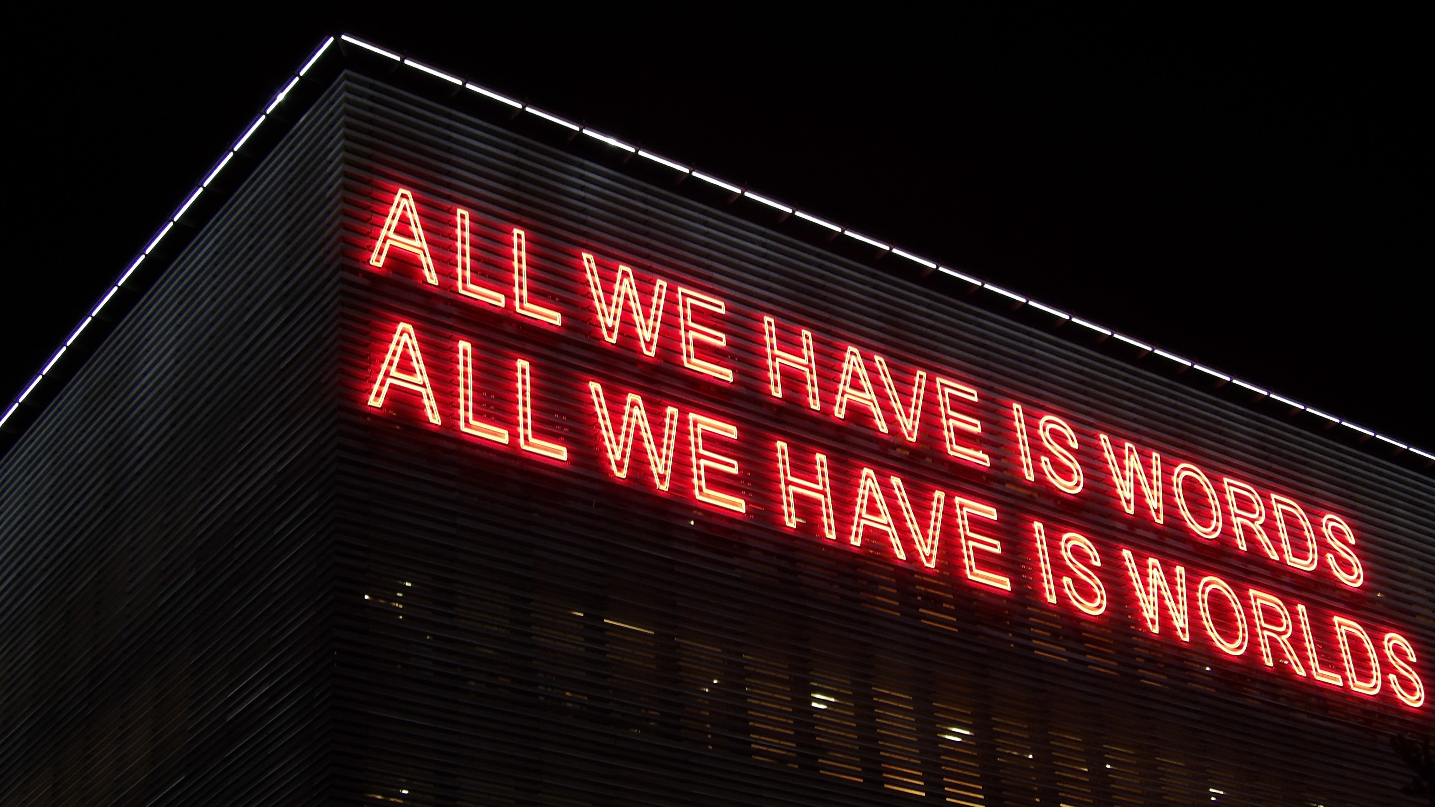 A large, red neon sign on the side of building that reads 'All We Have Is Words, All We Have Is Worlds"