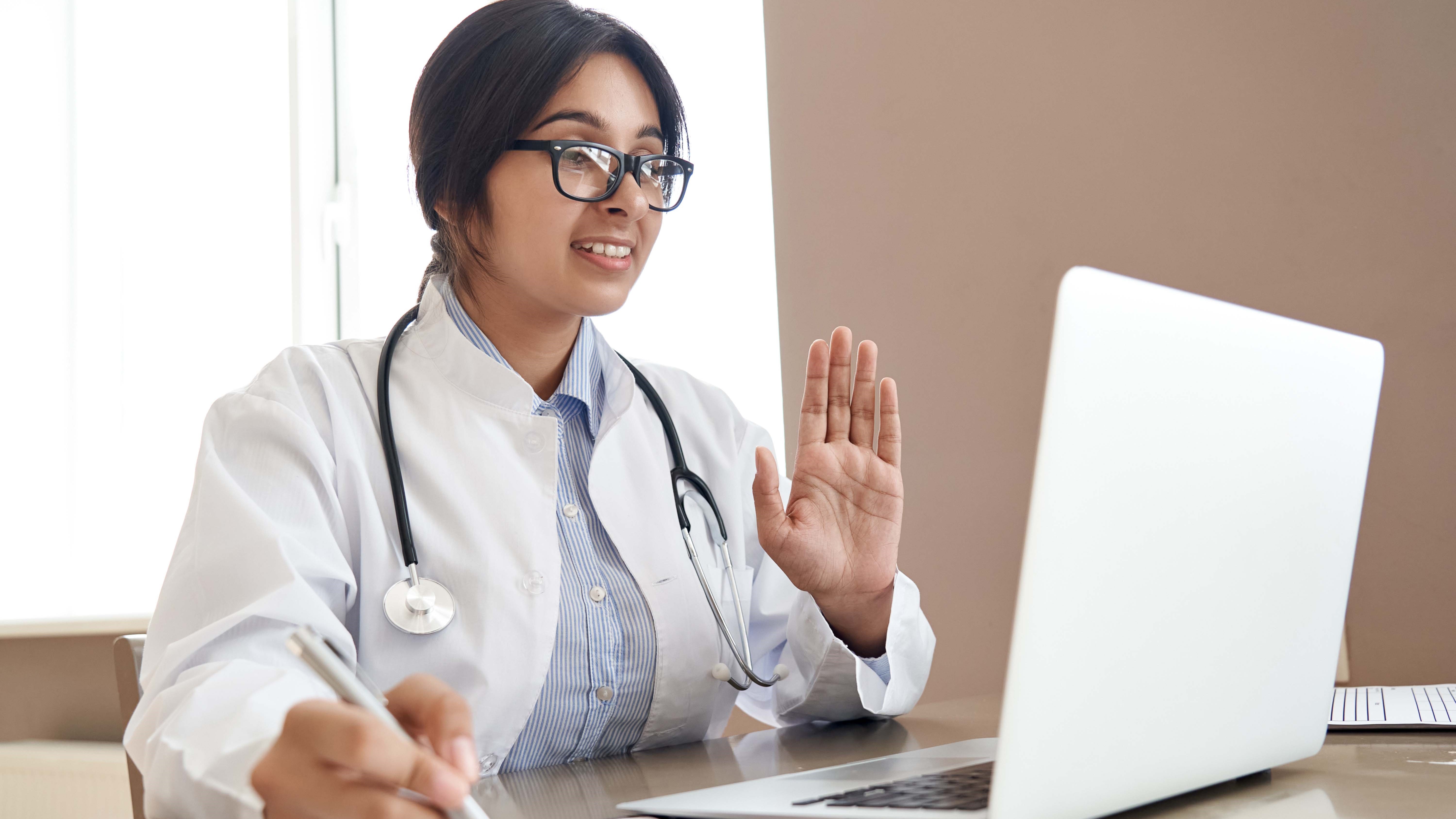 A person in a white coat and stethoscope has a video call on laptop