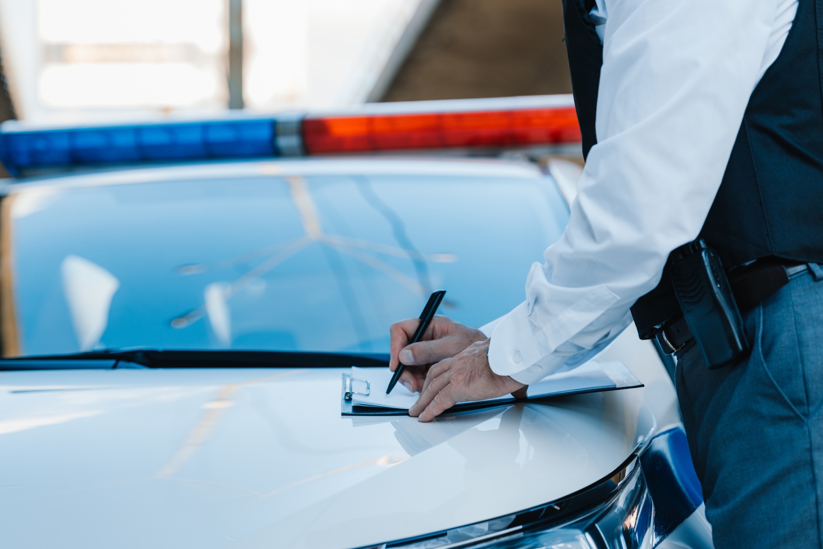 A police officer writes on a clipboard in front of a police car