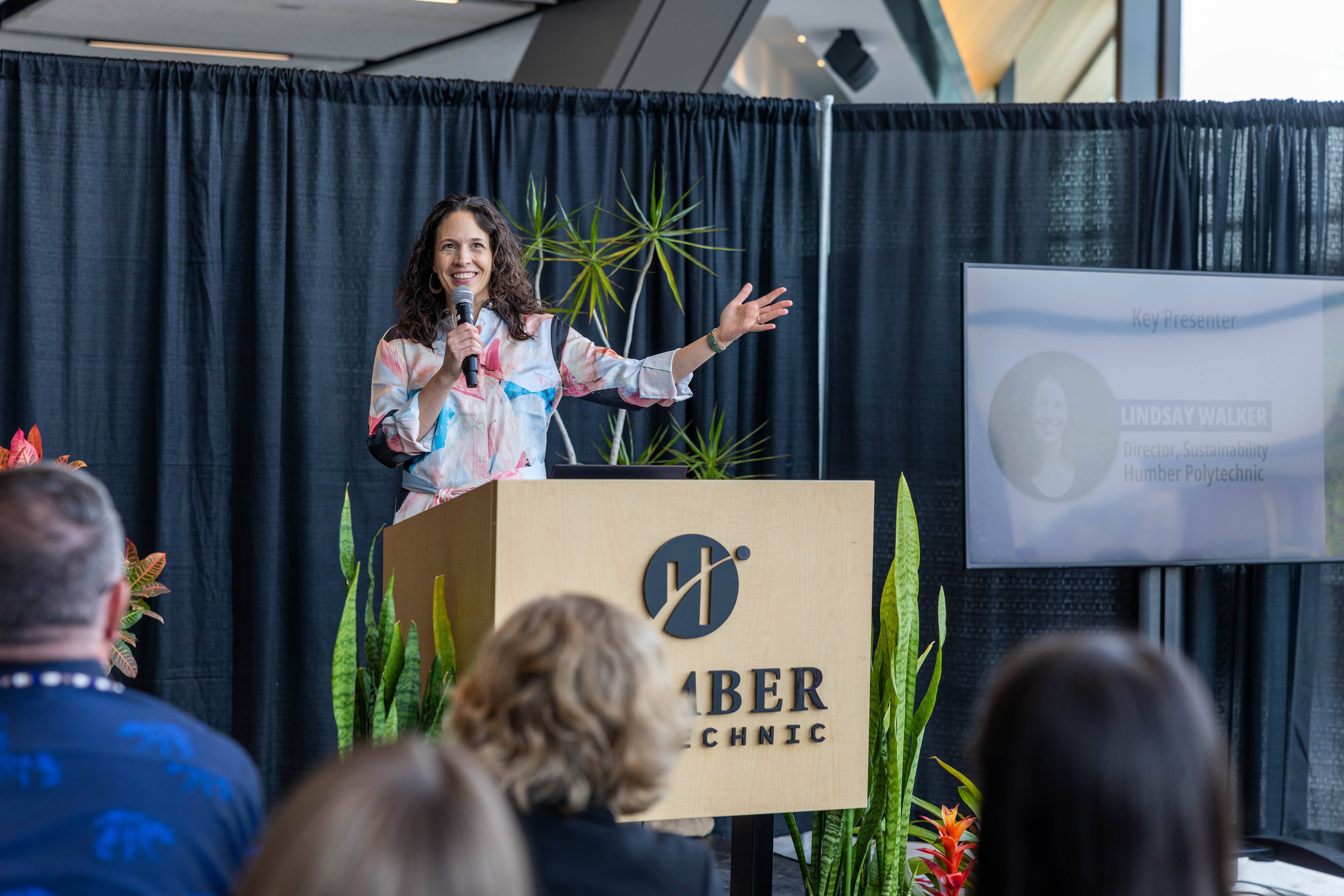 A woman stands at a podium surrounded by plants.